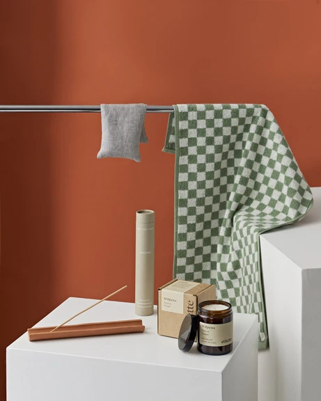 Still life of bath and home goods: towels, candle, incense. Checkered towel hanging on a silver towel rack against an orange wall.