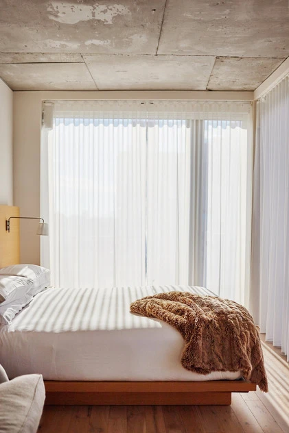 Bedroom interior featuring a queen-sized bed with white linens and a brown faux fur throw, bright natural light, and a concrete ceiling.