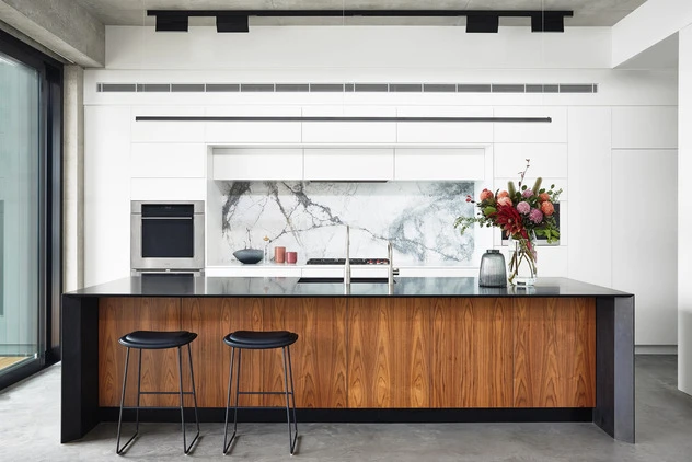 Modern kitchen interior featuring a wooden island with black countertop and two bar stools, set against a marble backsplash and white cabinetry.