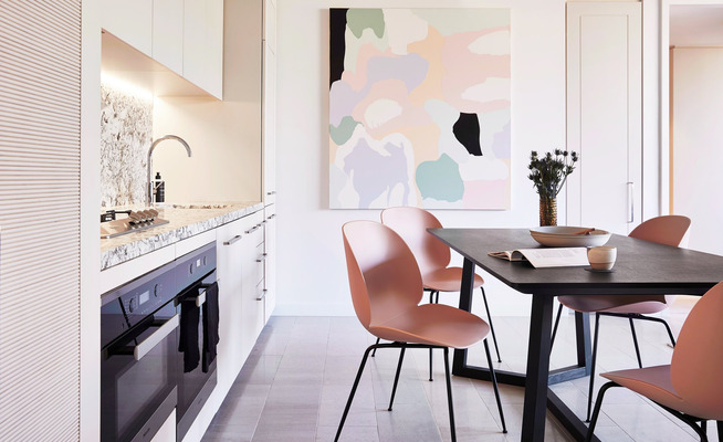 Interior shot of a modern kitchen and dining area with a black table, pink chairs, a white kitchen, and a marble countertop.