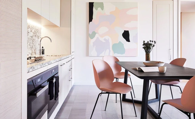 Interior shot of a modern kitchen and dining area with a black table, pink chairs, a white kitchen, and a marble countertop.