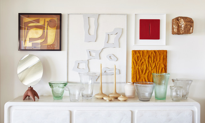Interior scene featuring a white credenza supporting vases, wooden sculptures, and framed geometric art, with a white wall background.