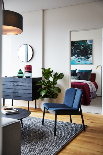 Contemporary living room with blue chair, modern dresser, potted plant, round mirror, and abstract artwork above bed.