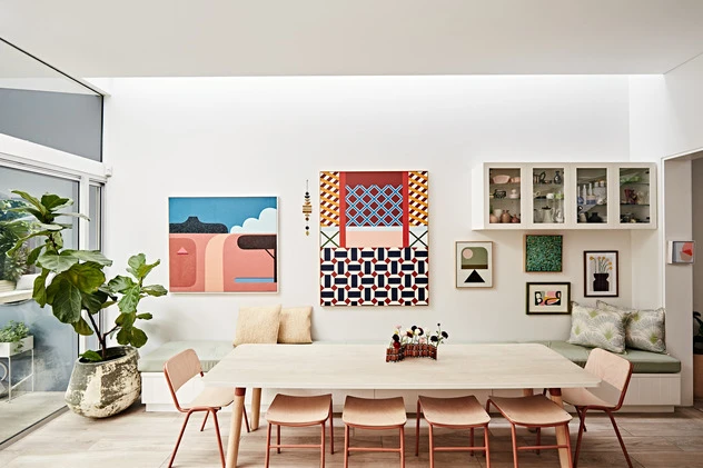 Well-lit modern dining room with a light wood table, pink chairs, and colorful artwork on the white wall, creating an inviting space.