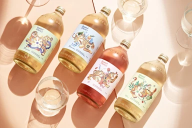 Four glass wine bottles with illustrated tiger labels and empty wine glasses arranged on a warm pink surface.