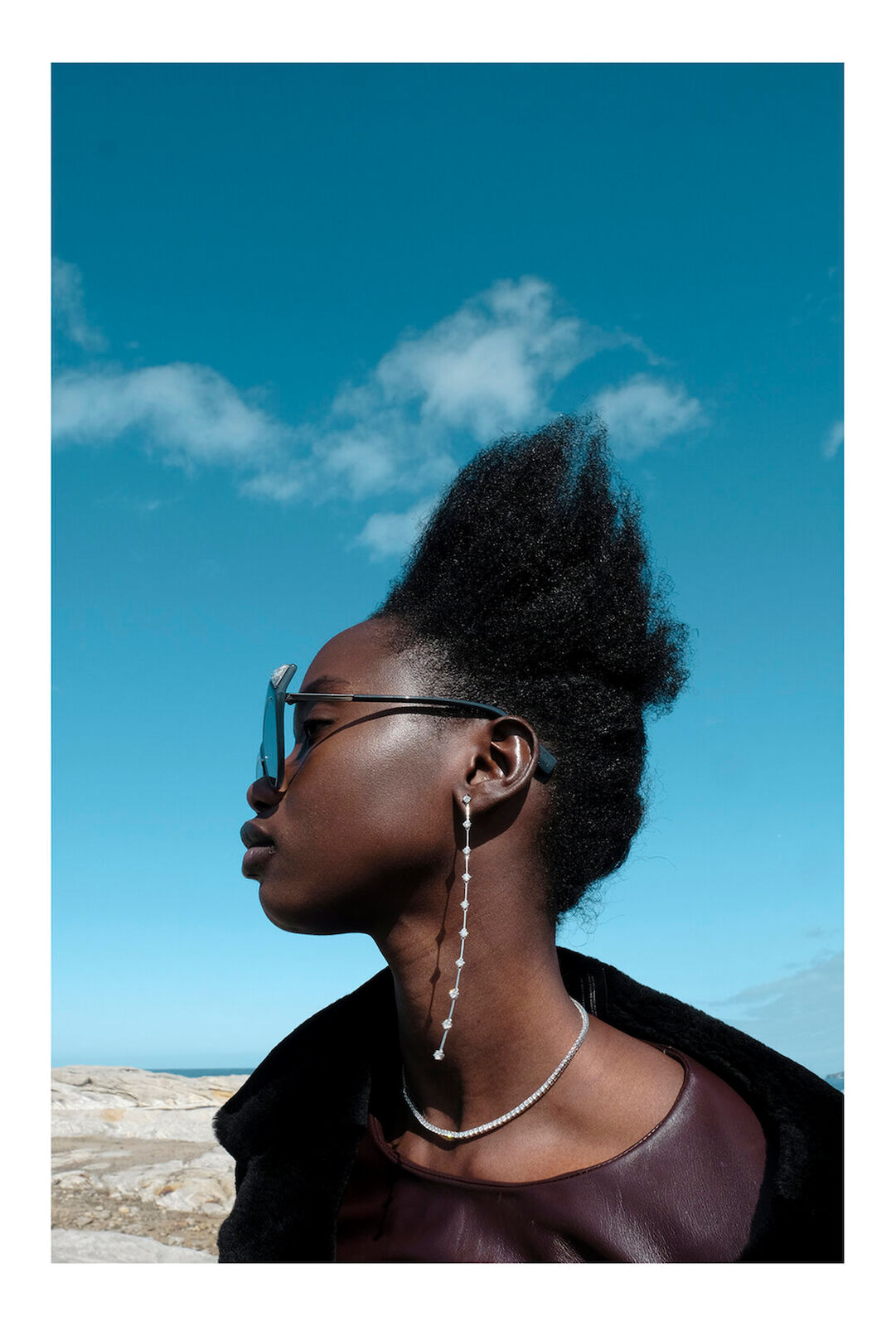 Eye-level portrait of an African woman with dark hair and sunglasses, posing against a blue sky with light clouds, wearing jewelry.