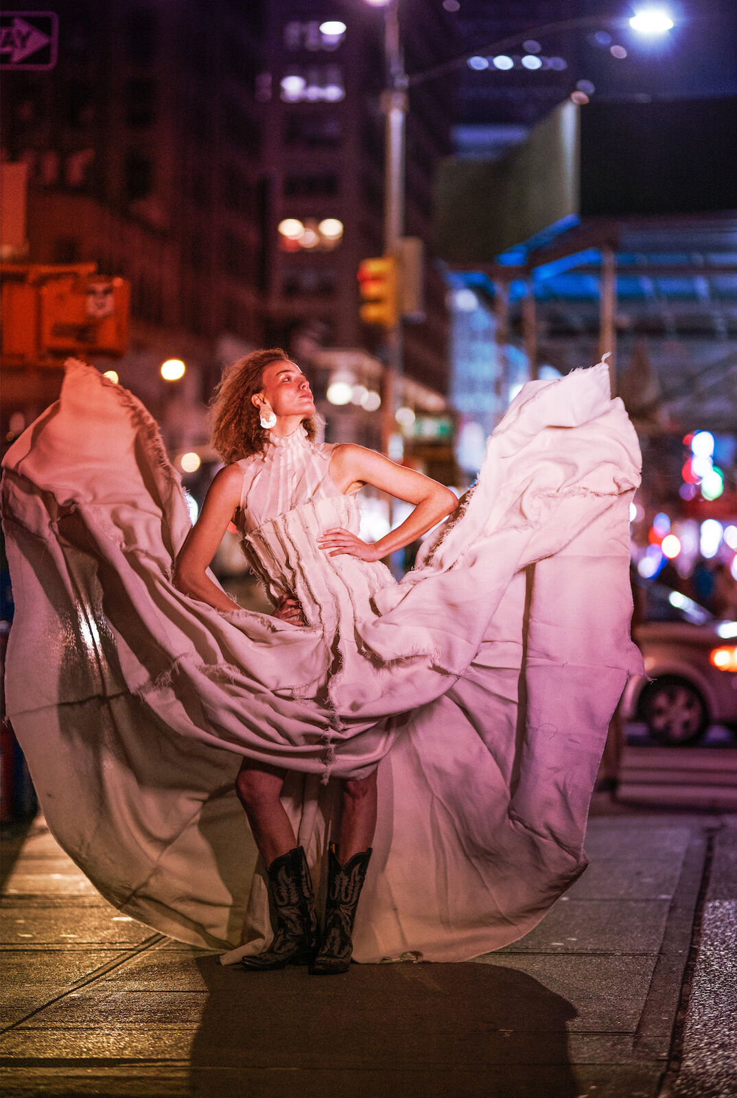 Fashion photography features a woman in a flowing dress and cowboy boots posing on a city street at night, under streetlights and traffic signals.