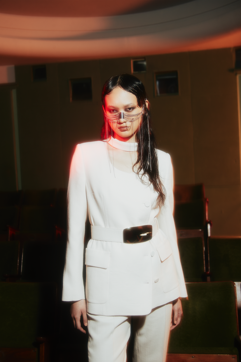 Fashion shot of a woman in a white suit with a face mask in a vintage theater setting with green seats, red lights, and dark tones.