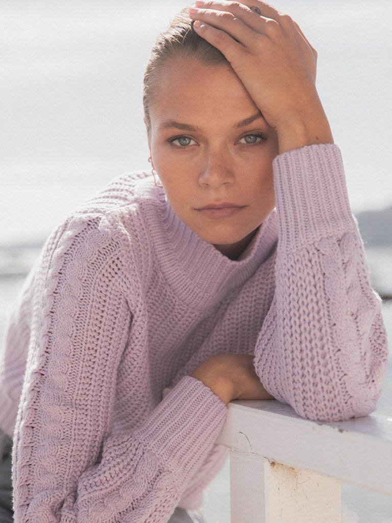 Portrait of a young woman with her hand on her head, wearing a light purple knitted sweater, leaning on a white railing outdoors.