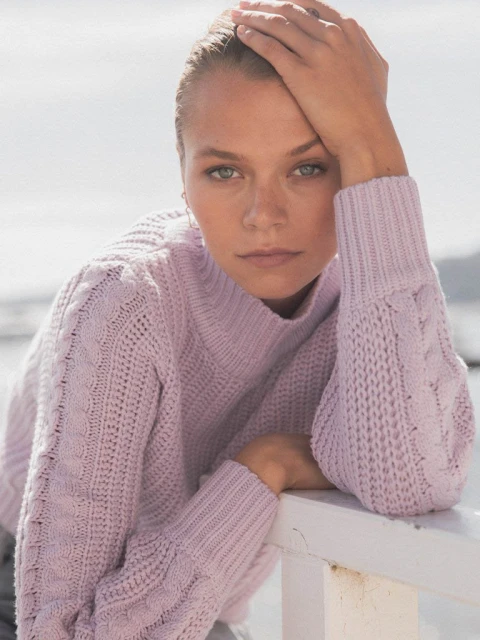 Portrait of a young woman with her hand on her head, wearing a light purple knitted sweater, leaning on a white railing outdoors.