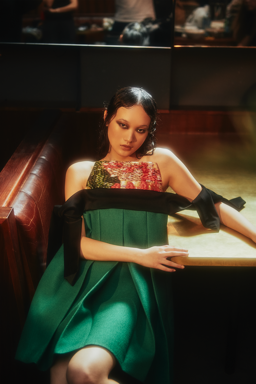 A woman in a green dress with floral accents poses indoors, seated at a table with moody, directional lighting casting shadows.