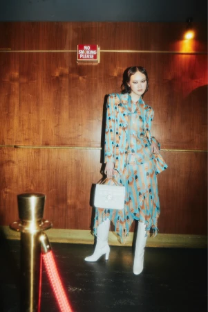 Woman in patterned blazer and skirt with white boots and purse poses in front of wood-paneled wall with 'No Smoking' sign.