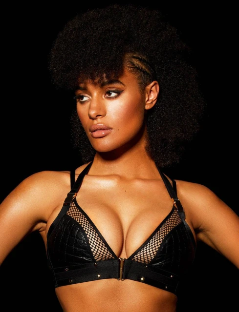 Portrait of a woman with dark curly hair, wearing a black bra with netting details against a dark background in a studio shot.