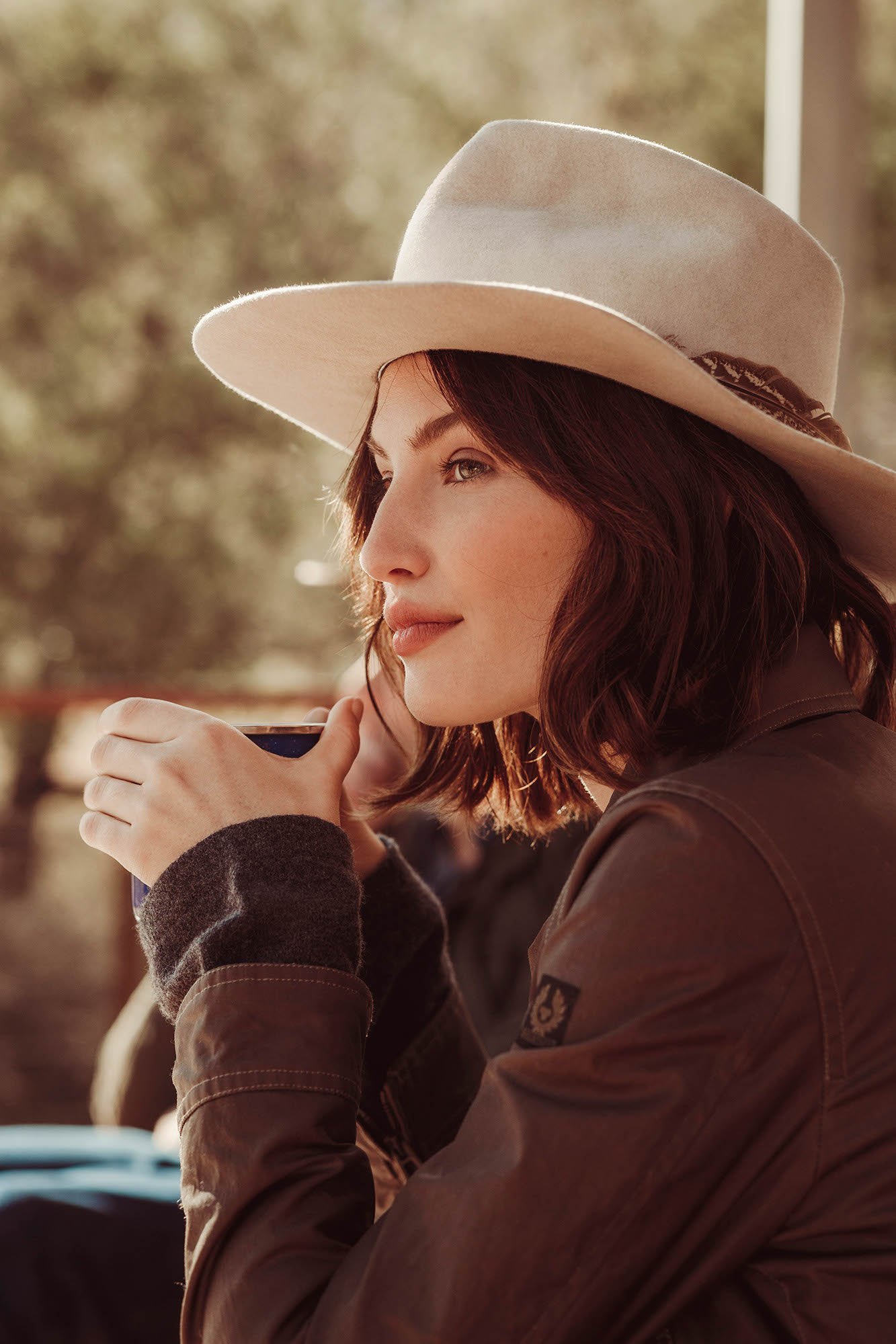Adult woman in cowboy hat sits outdoors, holding a cup. Brown jacket, short brown hair, serene expression, and diffused light.