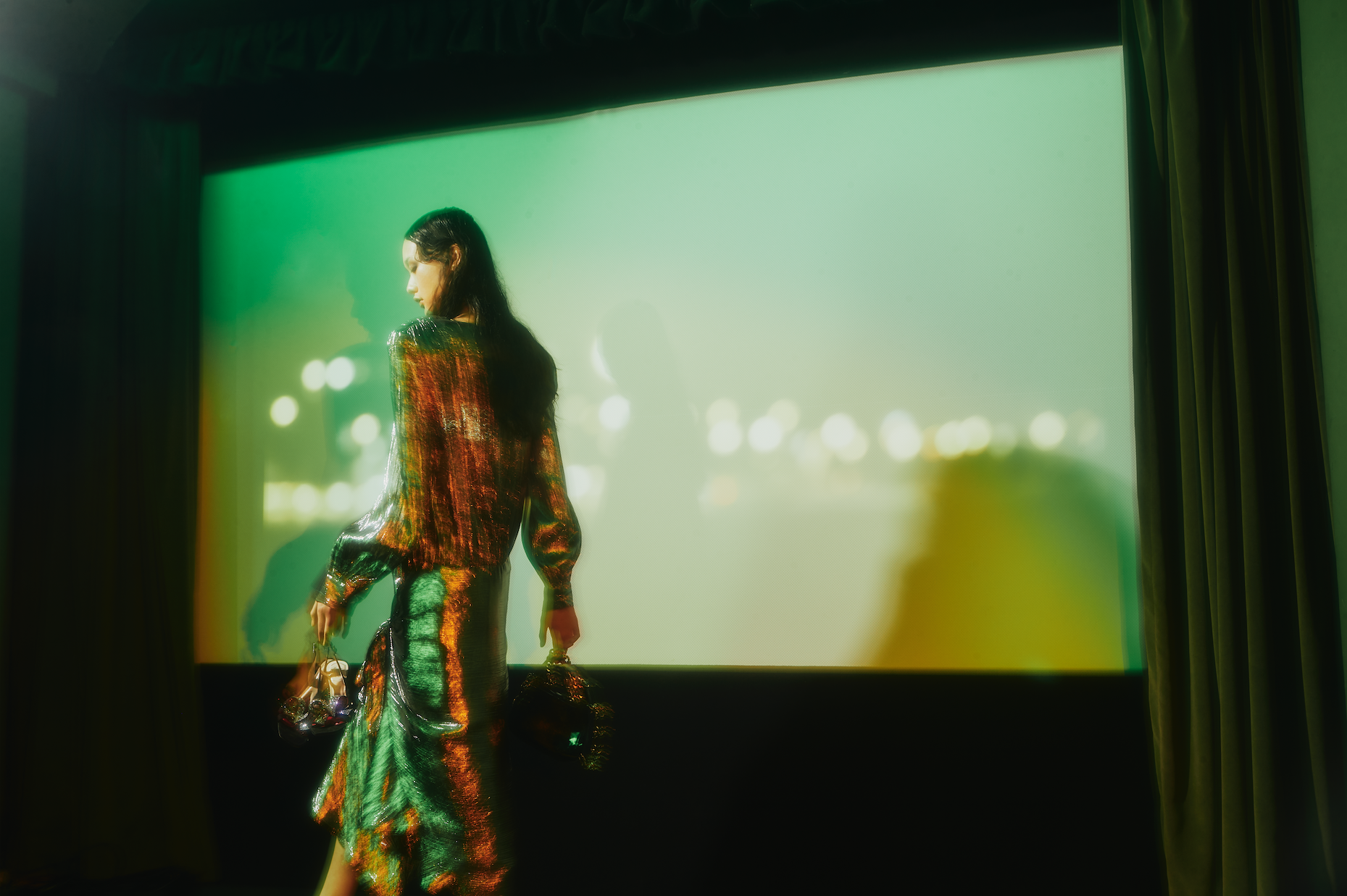 A woman in a green and orange sequin dress holds bags against a green screen with a blurred city lights background, fashion-forward.