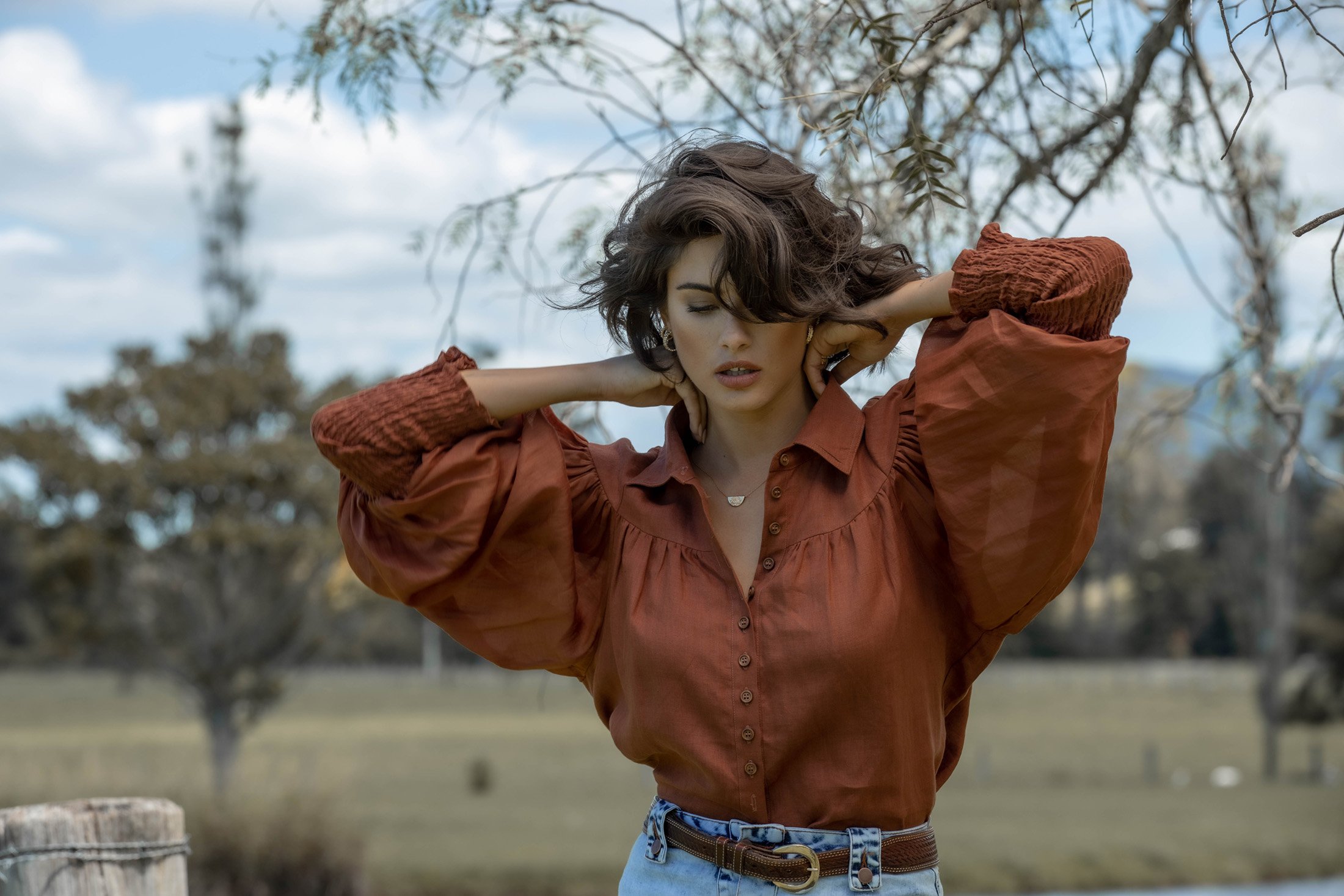 Brunette woman in brown blouse poses outdoors with hands behind her head against blurred background of trees, sky, and water.