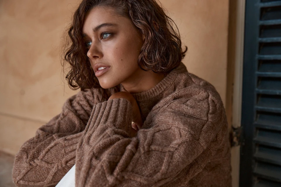 Close-up of a young adult woman with wavy brown hair wearing a brown cable knit sweater, looking off to the side with a neutral expression.
