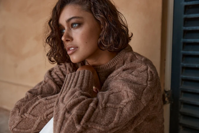 Close-up of a young adult woman with wavy brown hair wearing a brown cable knit sweater, looking off to the side with a neutral expression.