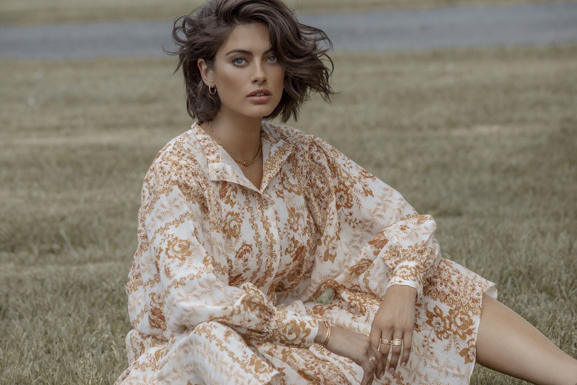 Woman in floral dress sits in a grassy field. She has short brown hair, fair skin, and is wearing gold jewelry in a natural outdoor setting.