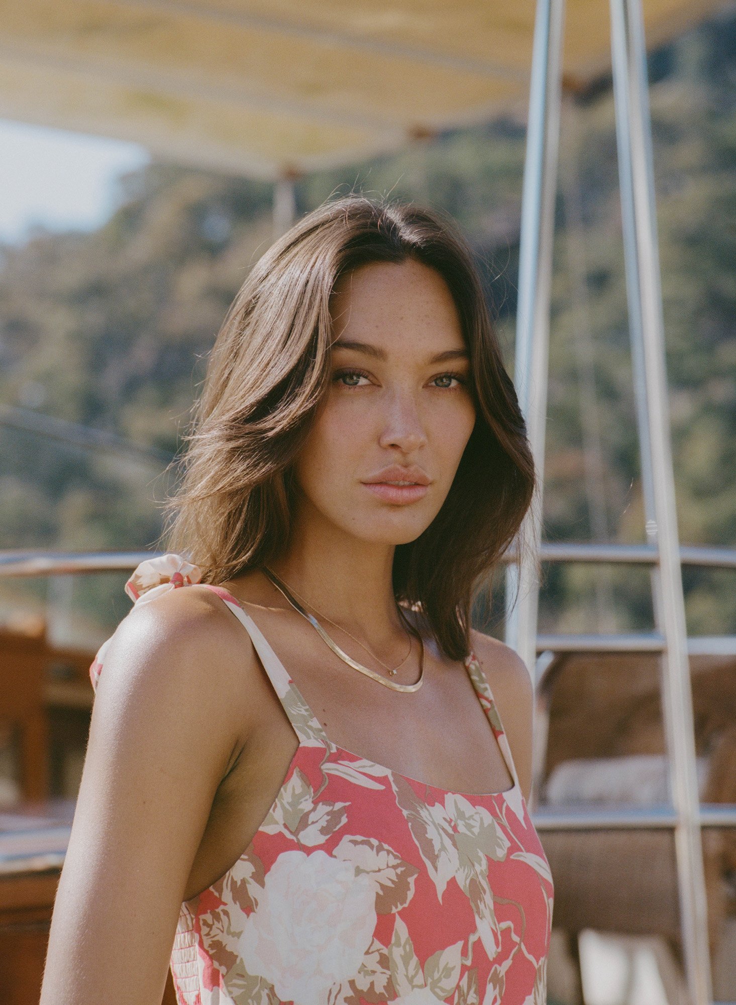 Portrait of a woman with brown hair wearing a floral dress. The background includes trees and a boat structure.
