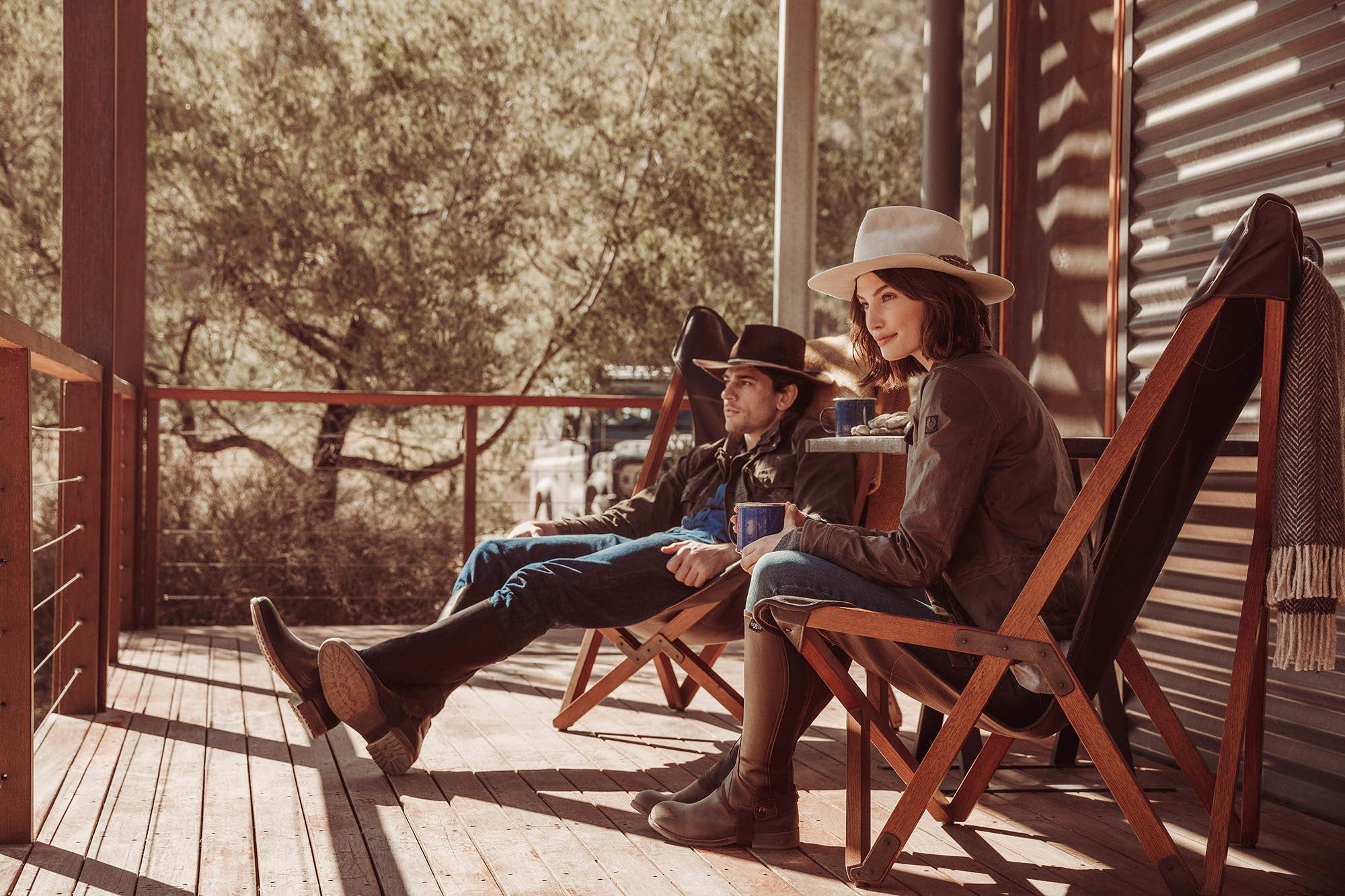 Man and woman wearing hats and boots seated on wooden folding chairs on a rustic timber deck in Sydney