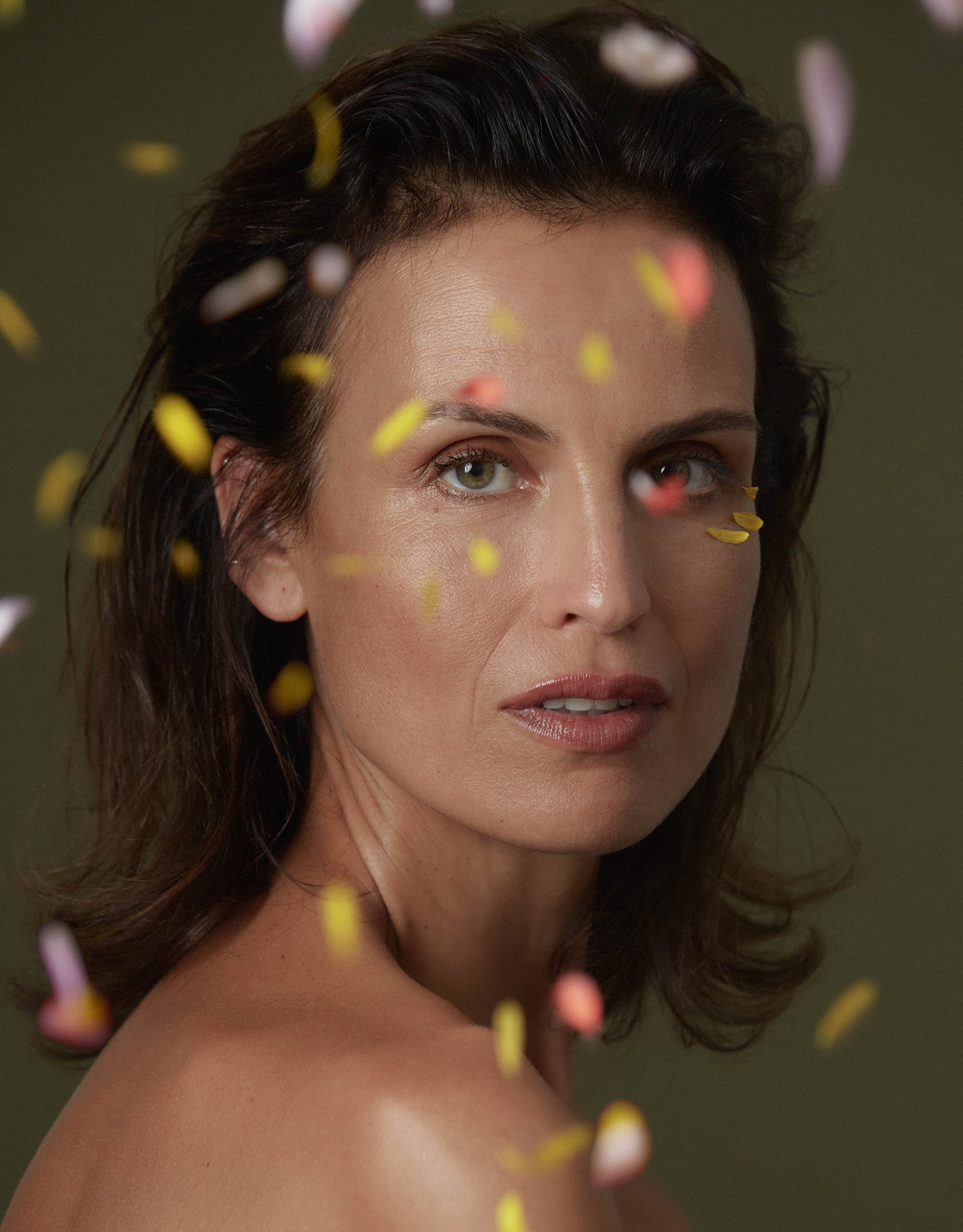 Portrait of a woman with bare shoulders and falling flower petals in a studio setting, featuring natural makeup and a contemplative expression.