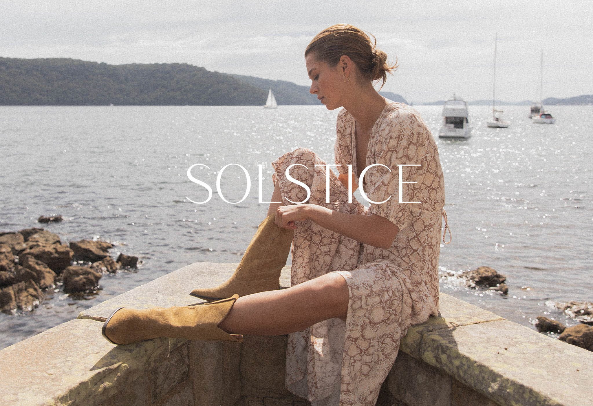 A woman in a snakeskin-patterned dress and tan suede boots sits on a stone wall by the ocean, sailboats visible on the water.