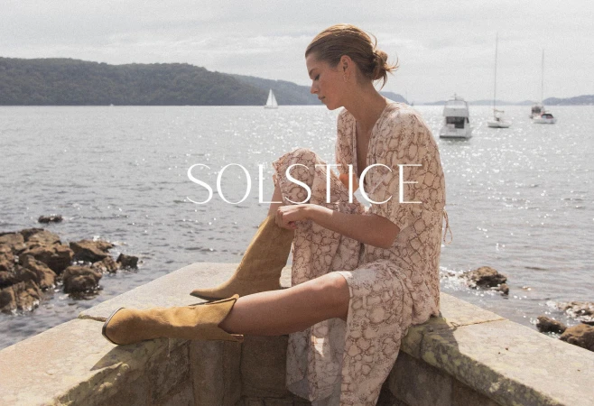 A woman in a snakeskin-patterned dress and tan suede boots sits on a stone wall by the ocean, sailboats visible on the water.
