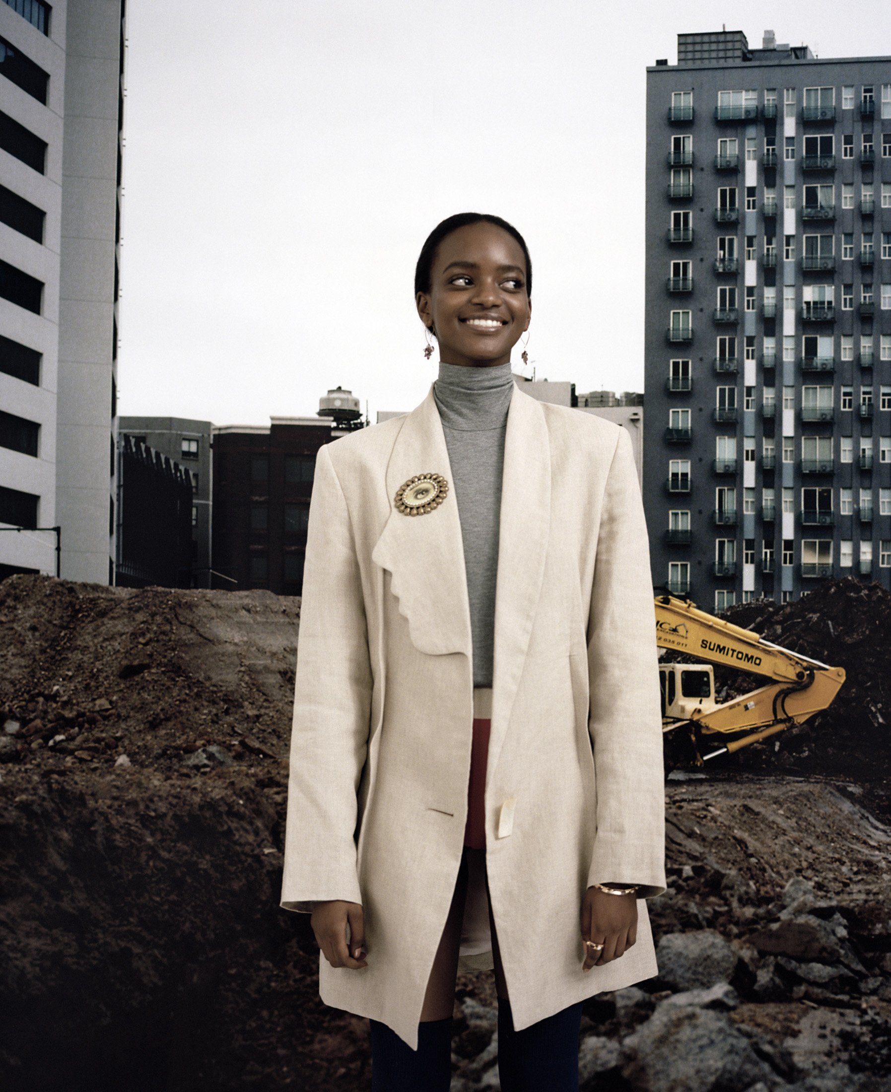 Woman in an off-white blazer poses against a backdrop of city buildings and a construction site with an excavator, looking away.