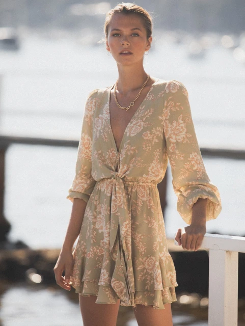 Woman in a floral print mini dress with long sleeves posing outdoors near a body of water and a retaining wall under bright sunlight.