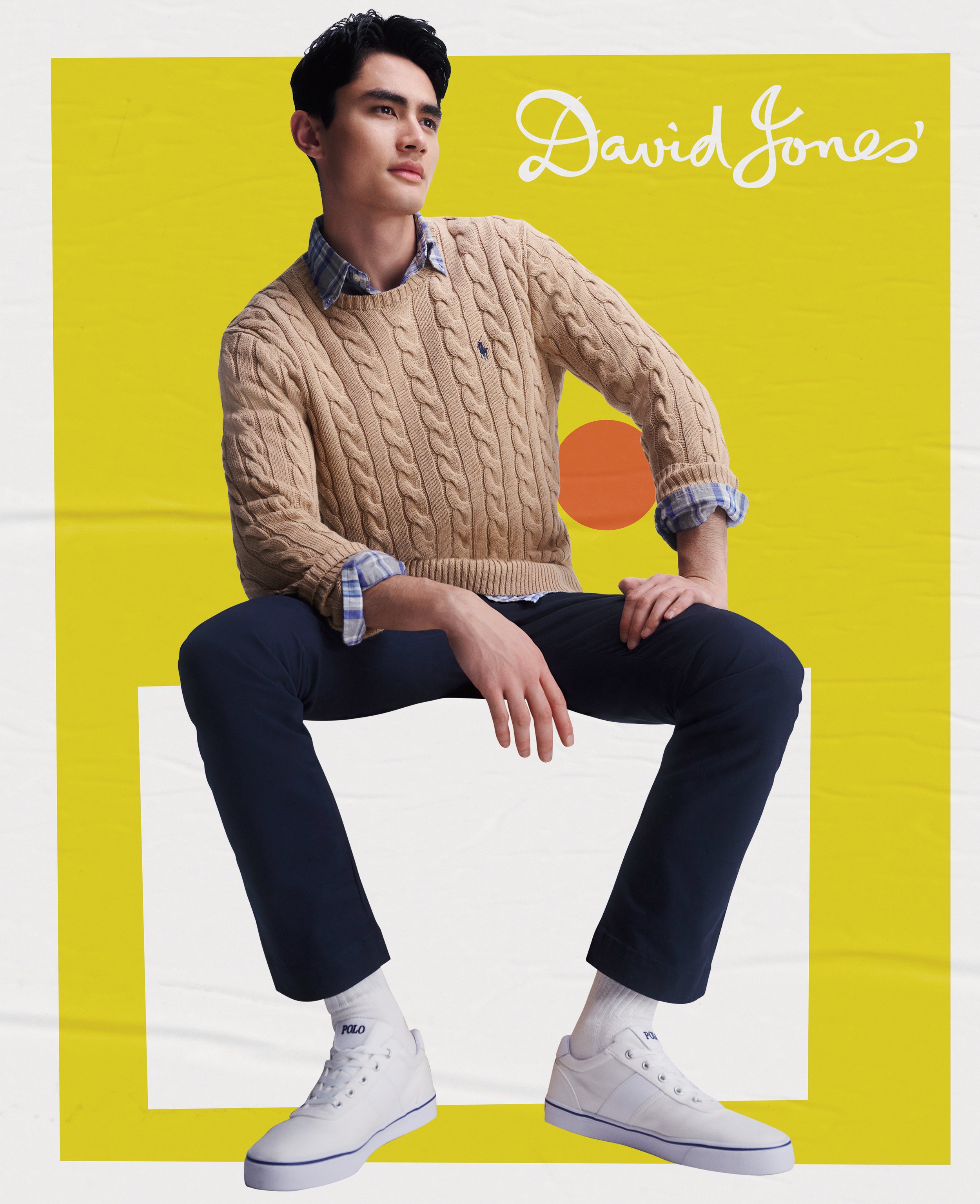 Man in cable-knit sweater seated indoors against a bold yellow backdrop wearing tailored navy pants and white sneakers, looking pensive.