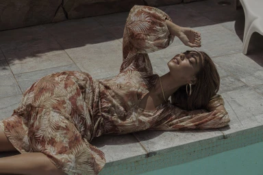 Woman wearing a brown patterned leaf-print dress reclining poolside with her arm raised above her head.