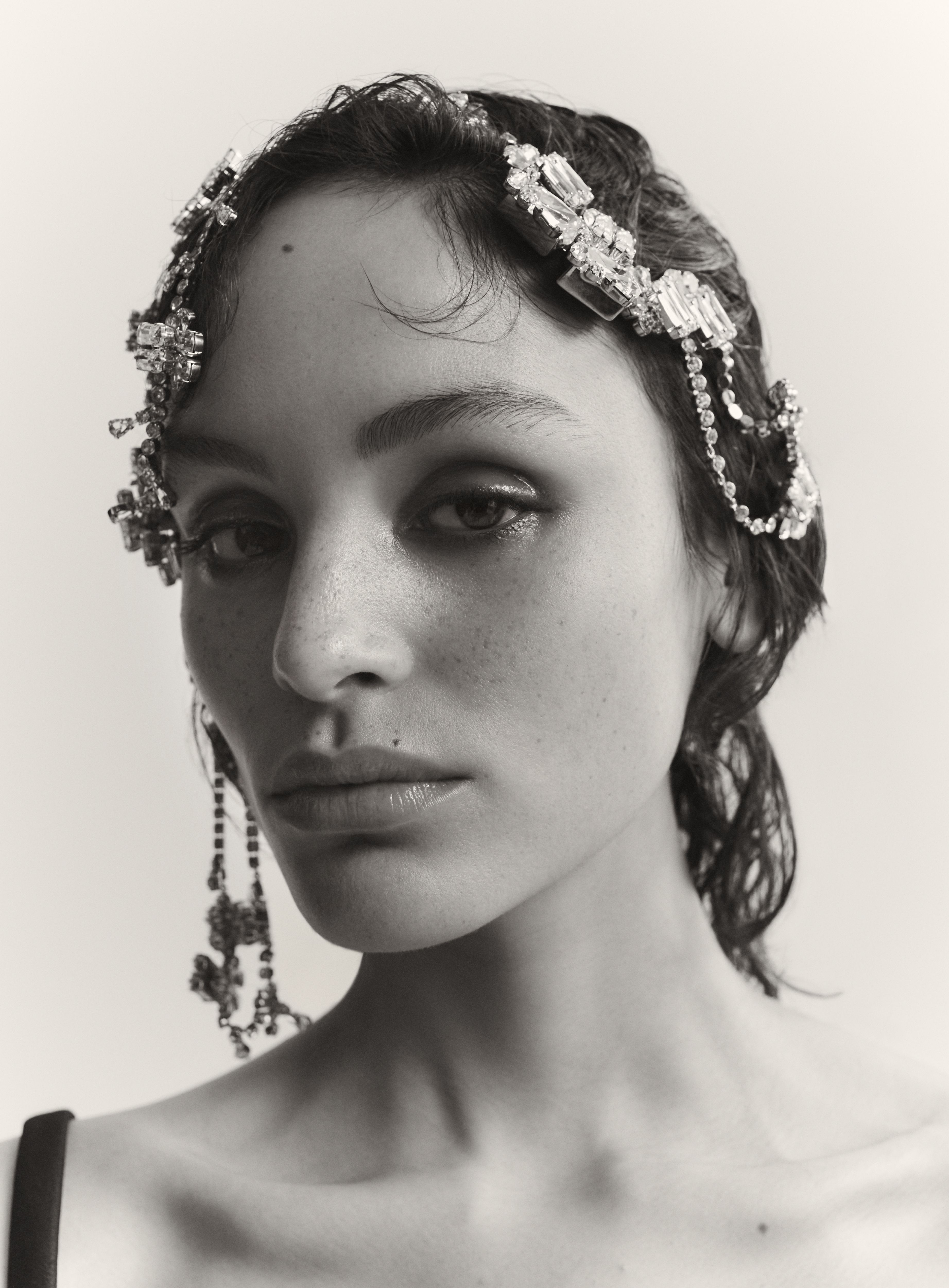 Close-up portrait of a woman with wet hair, wearing jeweled headwear and matching earrings against a neutral backdrop in black and white.