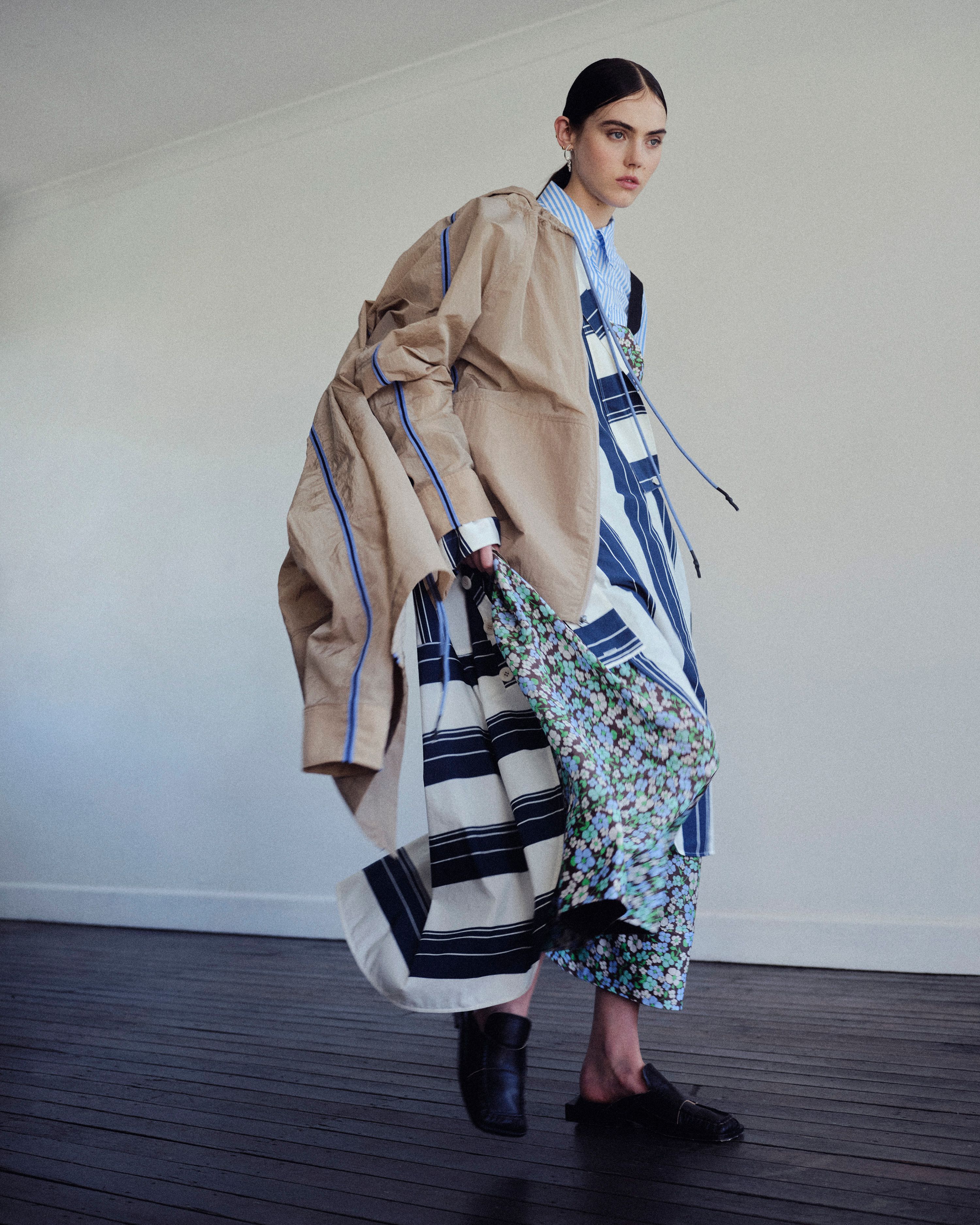 A fashionably dressed adult woman is walking indoors; she's wearing a striped shirt, khaki jacket, ankle boots, and patterned skirts.