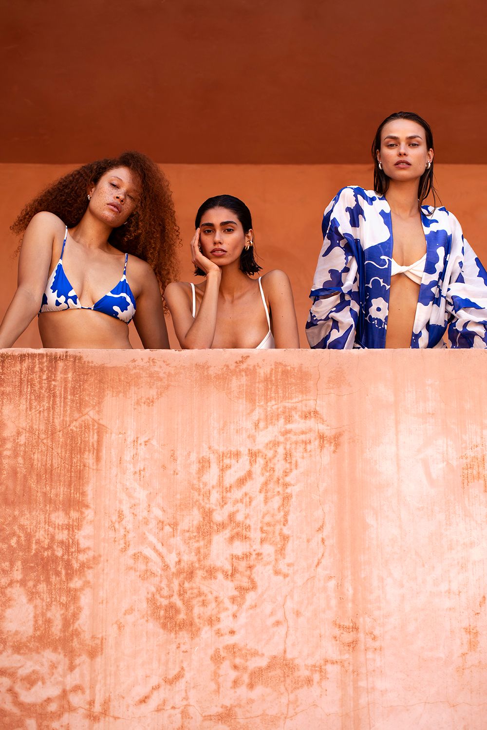 Three female models posing behind a terracotta ledge against a warm orange wall, styled with blue and white swimwear.