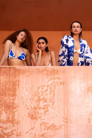 Three female models posing behind a terracotta ledge against a warm orange wall, styled with blue and white swimwear.