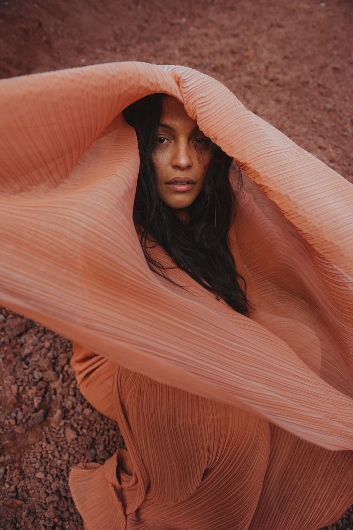 Portrait of a woman with long dark hair, draped in a peach-colored pleated fabric against a textured red-brown earth backdrop.