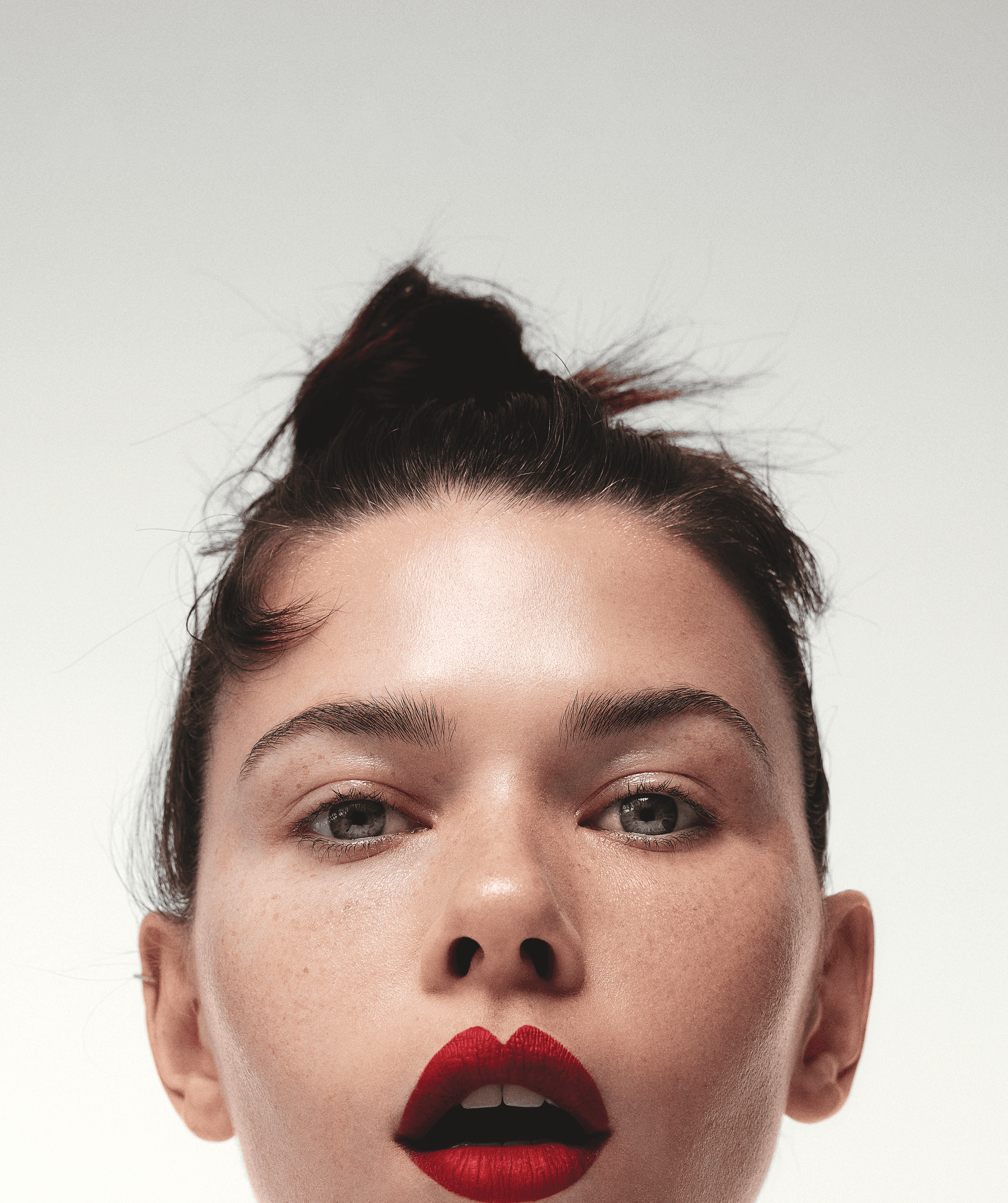 Close-up portrait of a young adult female with a topknot hairstyle and bright red lipstick against a plain, light background.