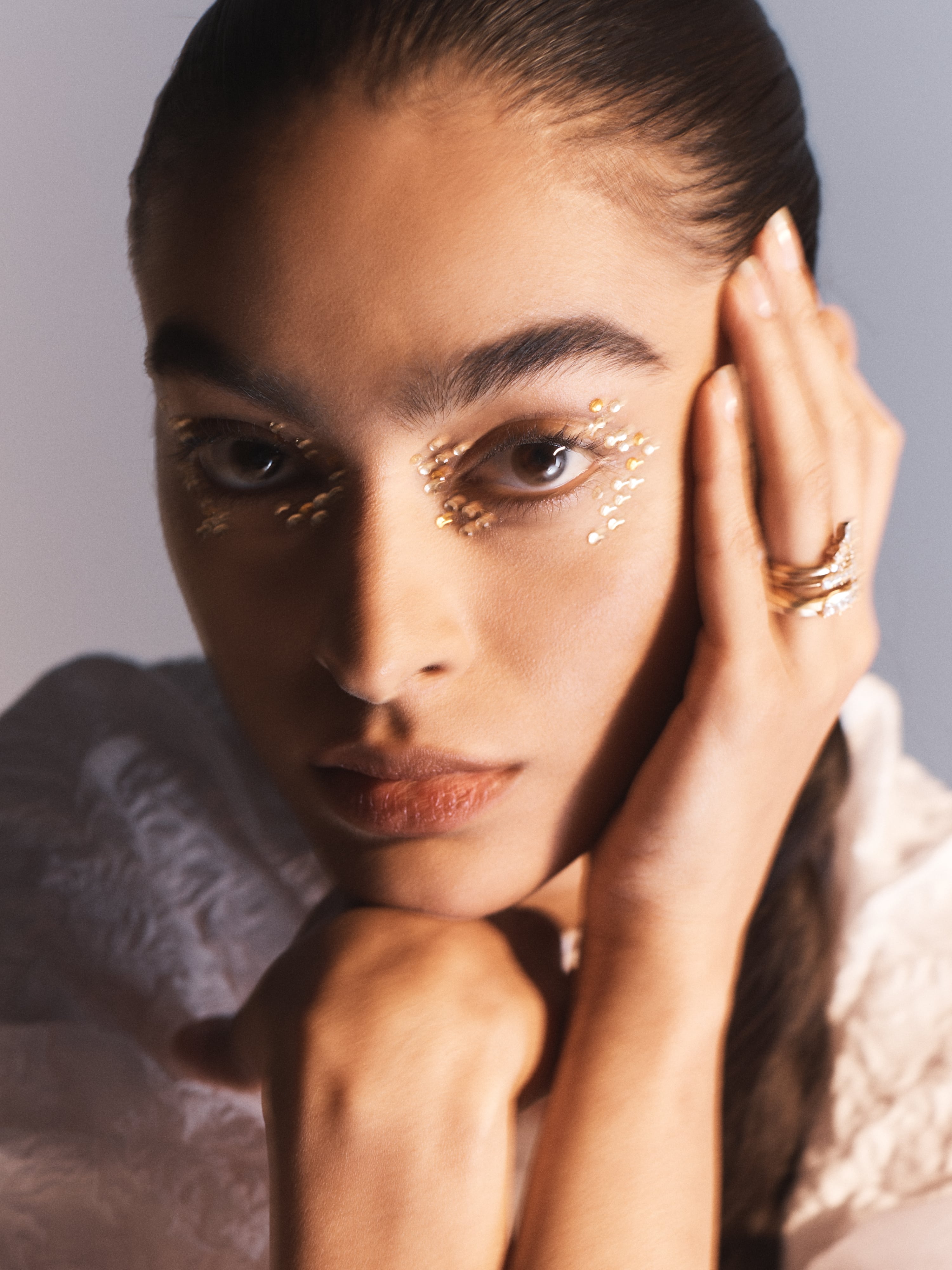Close-up of a beautiful adult female model with gold glitter makeup around her eyes and gold rings on her fingers against a grey background.