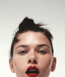 Close-up beauty portrait of a woman with vibrant red lipstick and hair in an updo against a neutral background.