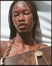 Close-up portrait of a Black woman with braided hair wearing gold hoop earrings and holding a brown monogram handbag with a chunky chain strap