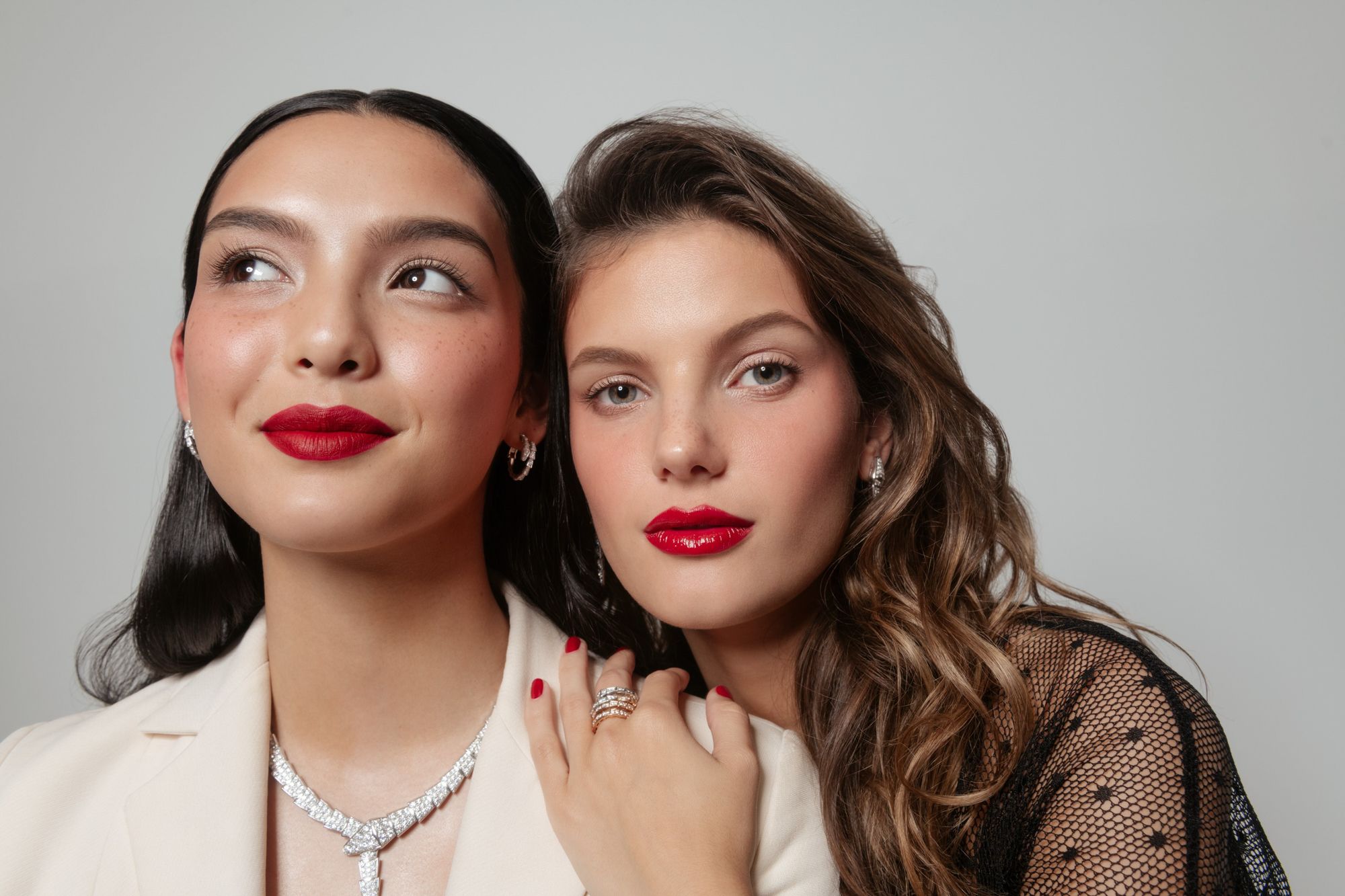 Two women pose in a beauty shot, wearing jewelry and red lipstick, against a neutral background. Shoulder-up view with a soft, diffused light.
