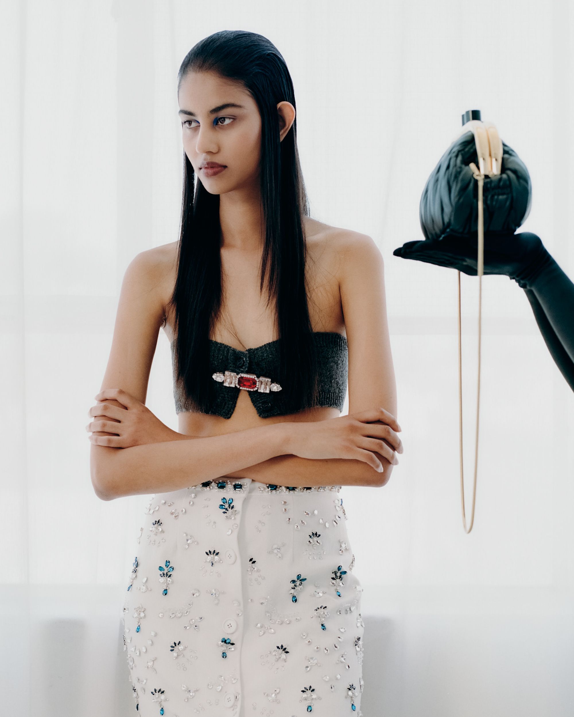 Woman in a stylish crop top and skirt holds a black purse with gold details, showcased in a bright, high-key setting.