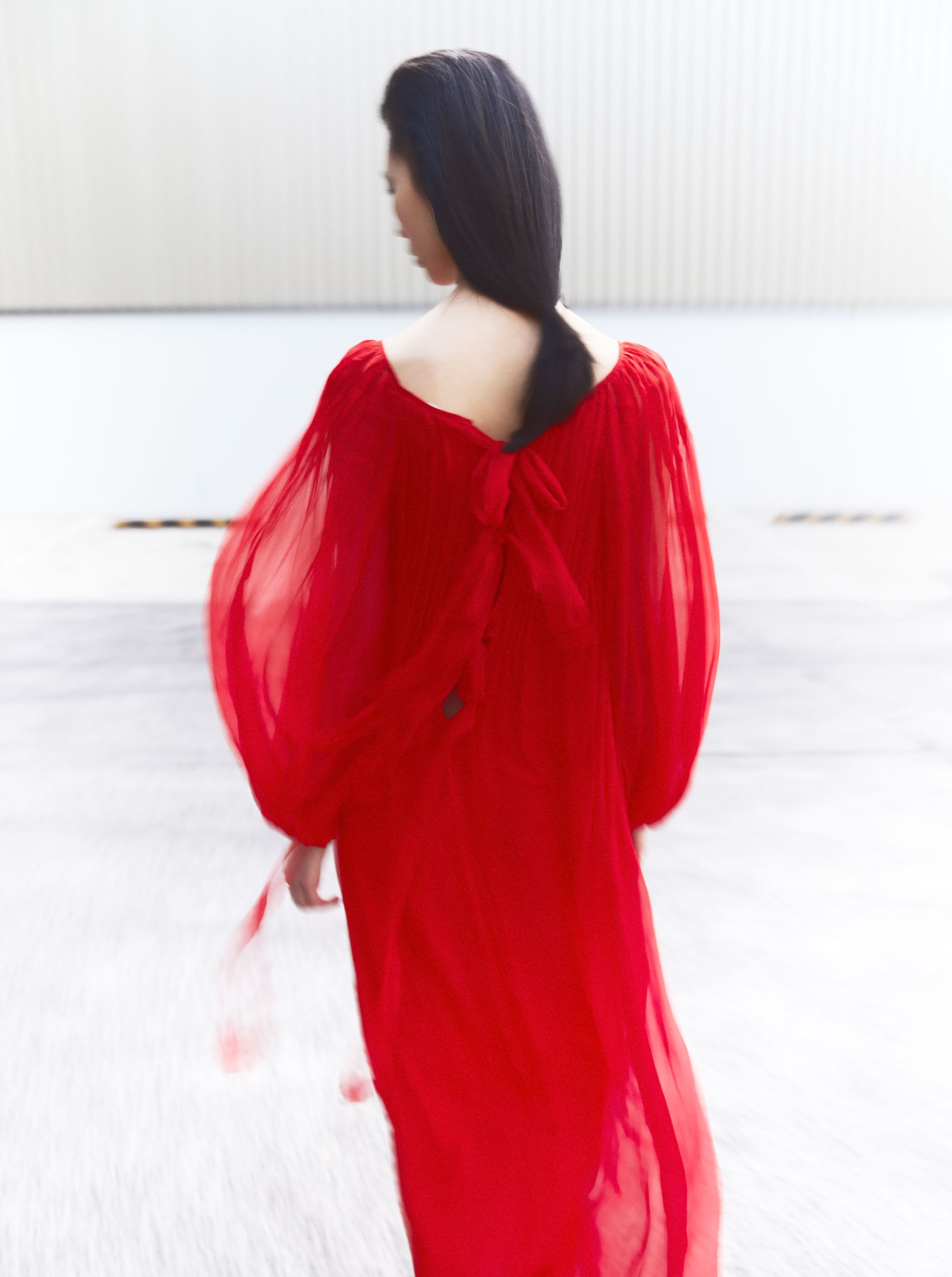 A woman with dark hair is seen from the back wearing a flowing red gown against a neutral industrial background, creating a striking contrast.