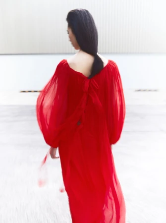 A woman with dark hair is seen from the back wearing a flowing red gown against a neutral industrial background, creating a striking contrast.