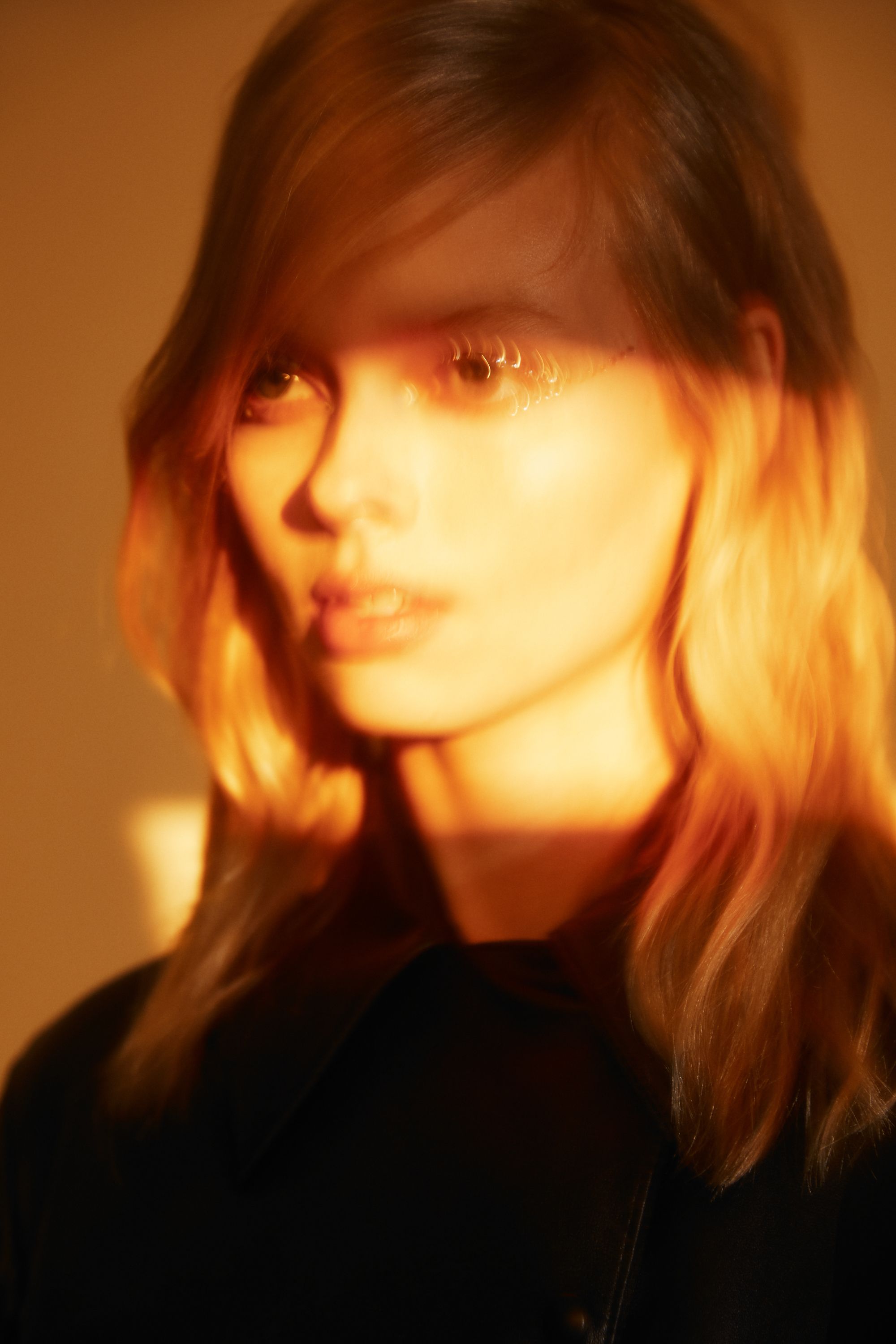 Close-up portrait of a young woman with blonde hair, wearing a black jacket and illuminated by warm golden light.