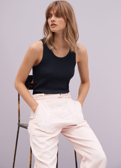 Model with long blonde hair wearing a black tank top and pale pink trousers, seated on a chair against a plain grey backdrop.