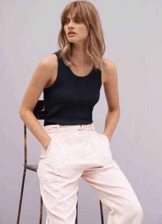 Model with long blonde hair wearing a black tank top and pale pink trousers, seated on a chair against a plain grey backdrop.