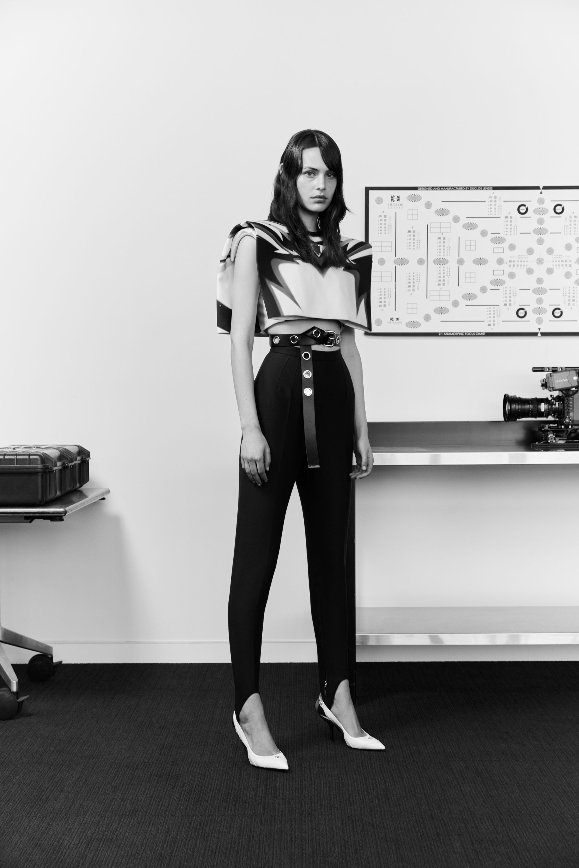 Studio shot of a young adult female fashion model in a black and white outfit, standing in front of a minimalist white background.