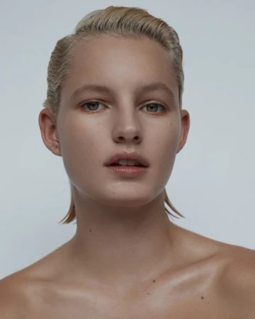 Close-up portrait of a woman with slicked-back blonde hair and natural makeup against a plain grey background